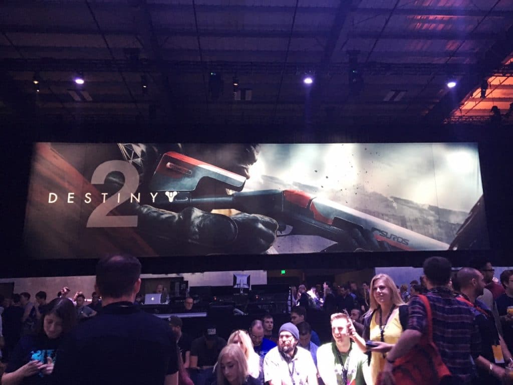 Inside photo of Destiny 2 event, crowd of people standing in front of large banner that says "Destiny 2" on it with a man holding a shotgun branded with Suros.